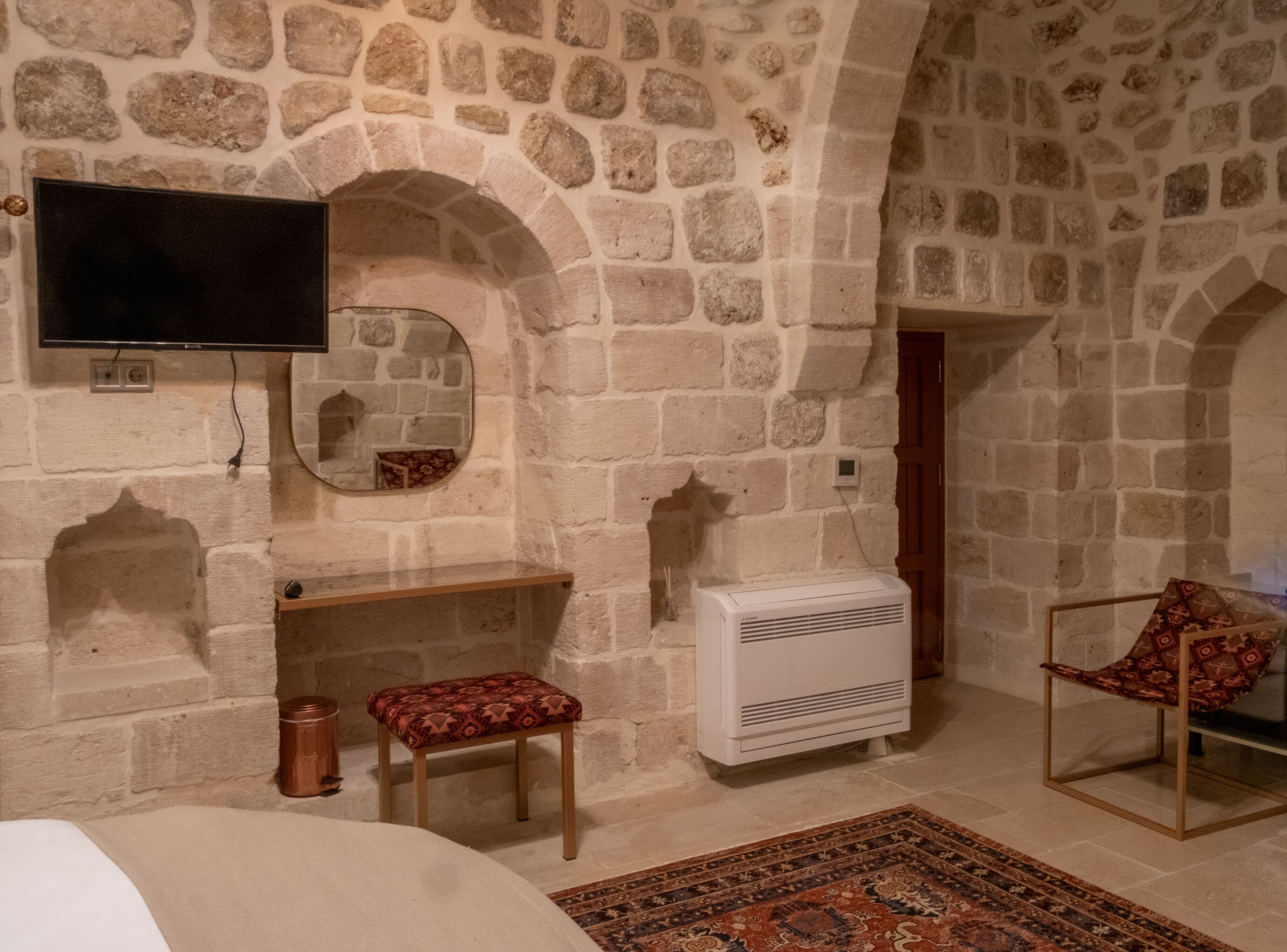 A stone-walled room with a flat-screen TV mounted above a wooden bench, a small table, a radiator, and a patterned rug on the floor.