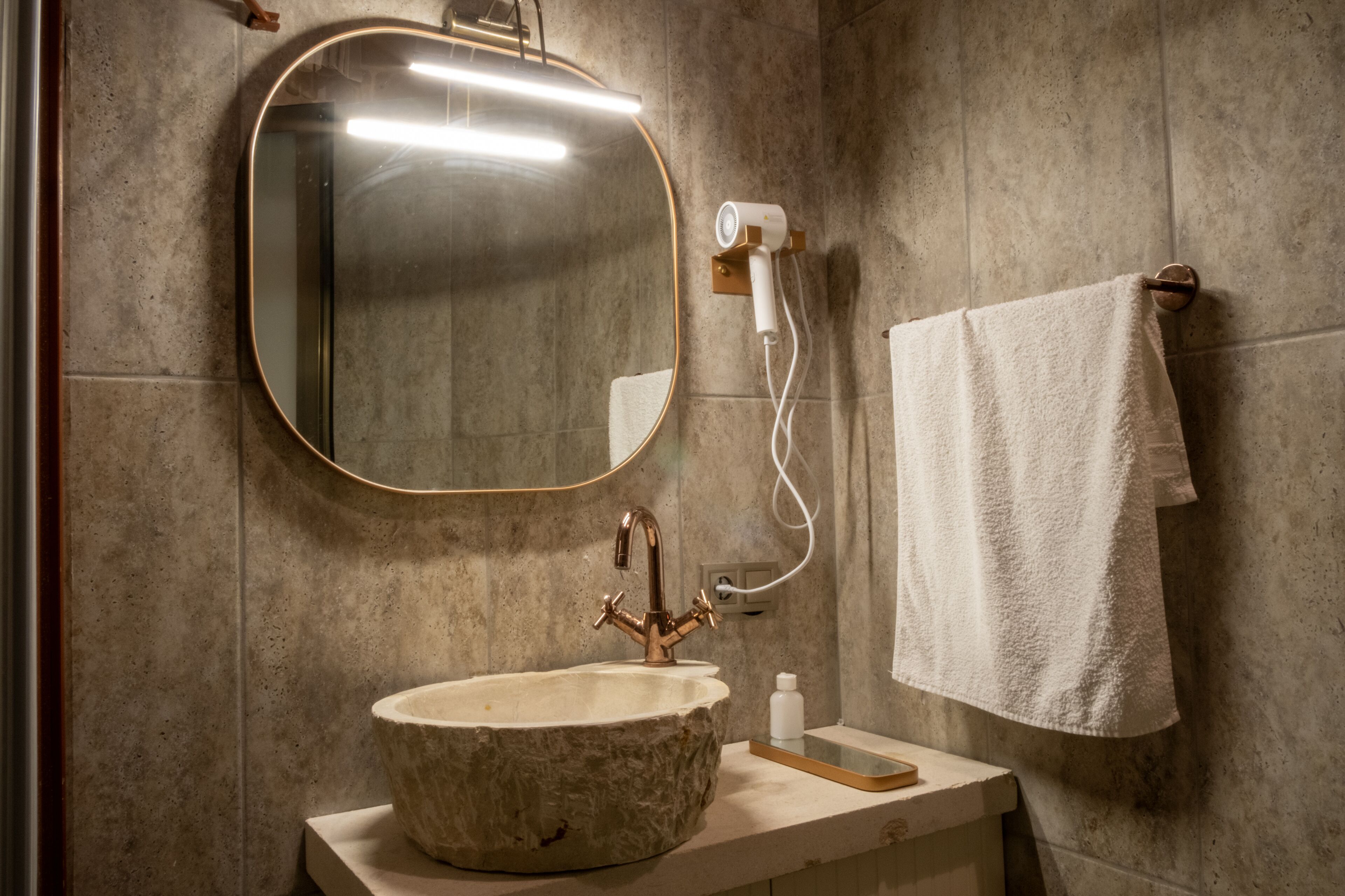 Un baño con lavabo de piedra, un espejo redondo, una toalla colgada en un perchero y un secador de pelo en la pared.