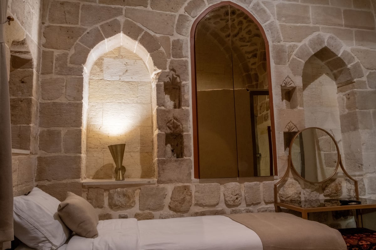 A stone-walled room with a bed, a mirror, and a window with a light fixture.