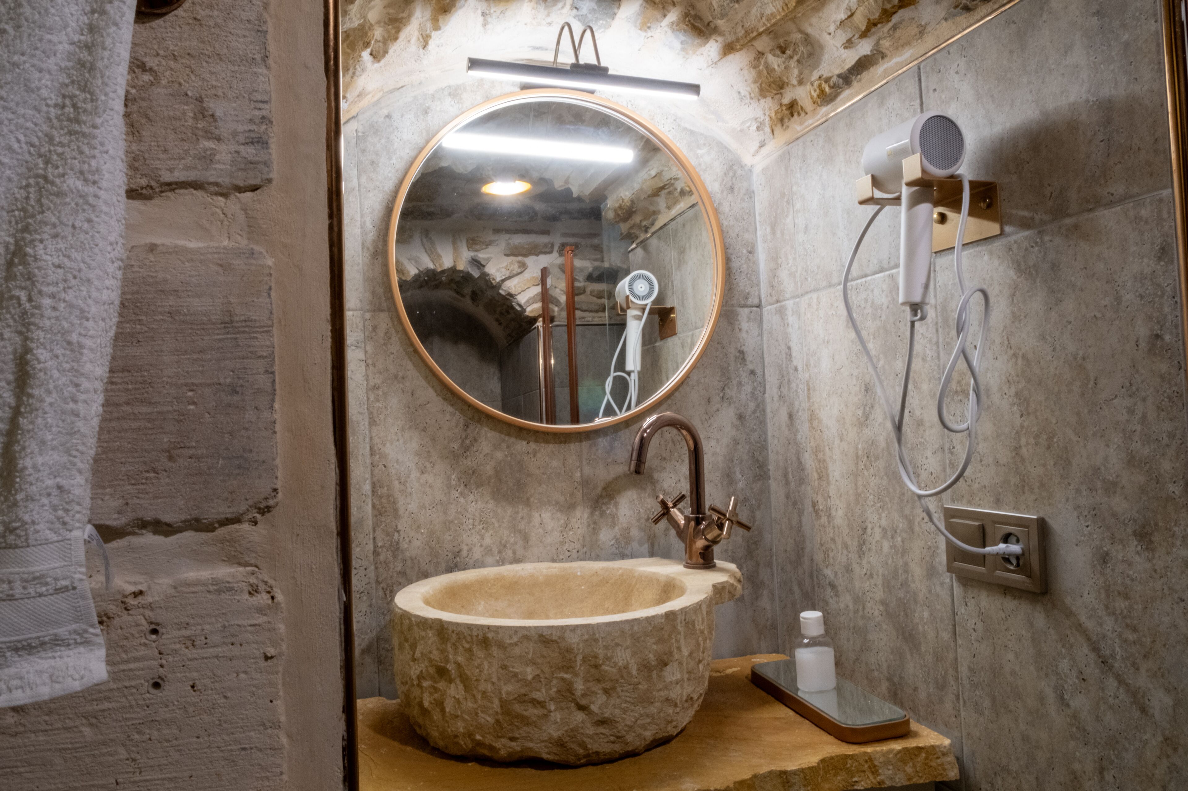 Une salle de bain avec un lavabo en pierre, un miroir rond, une douchette à main et un porte-serviettes.