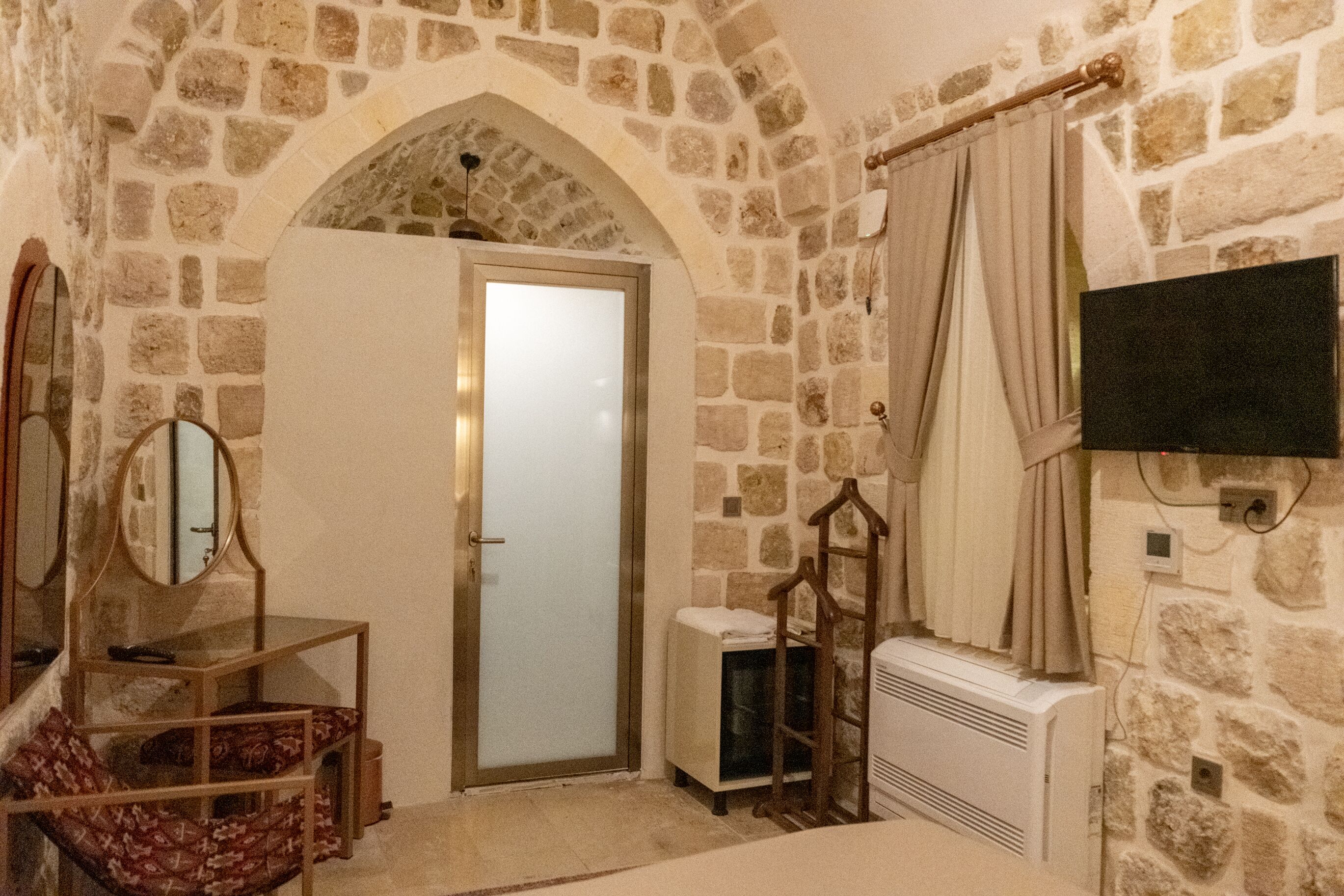 A stone-walled room with a door, a mirror, a radiator, and a television mounted on the wall.