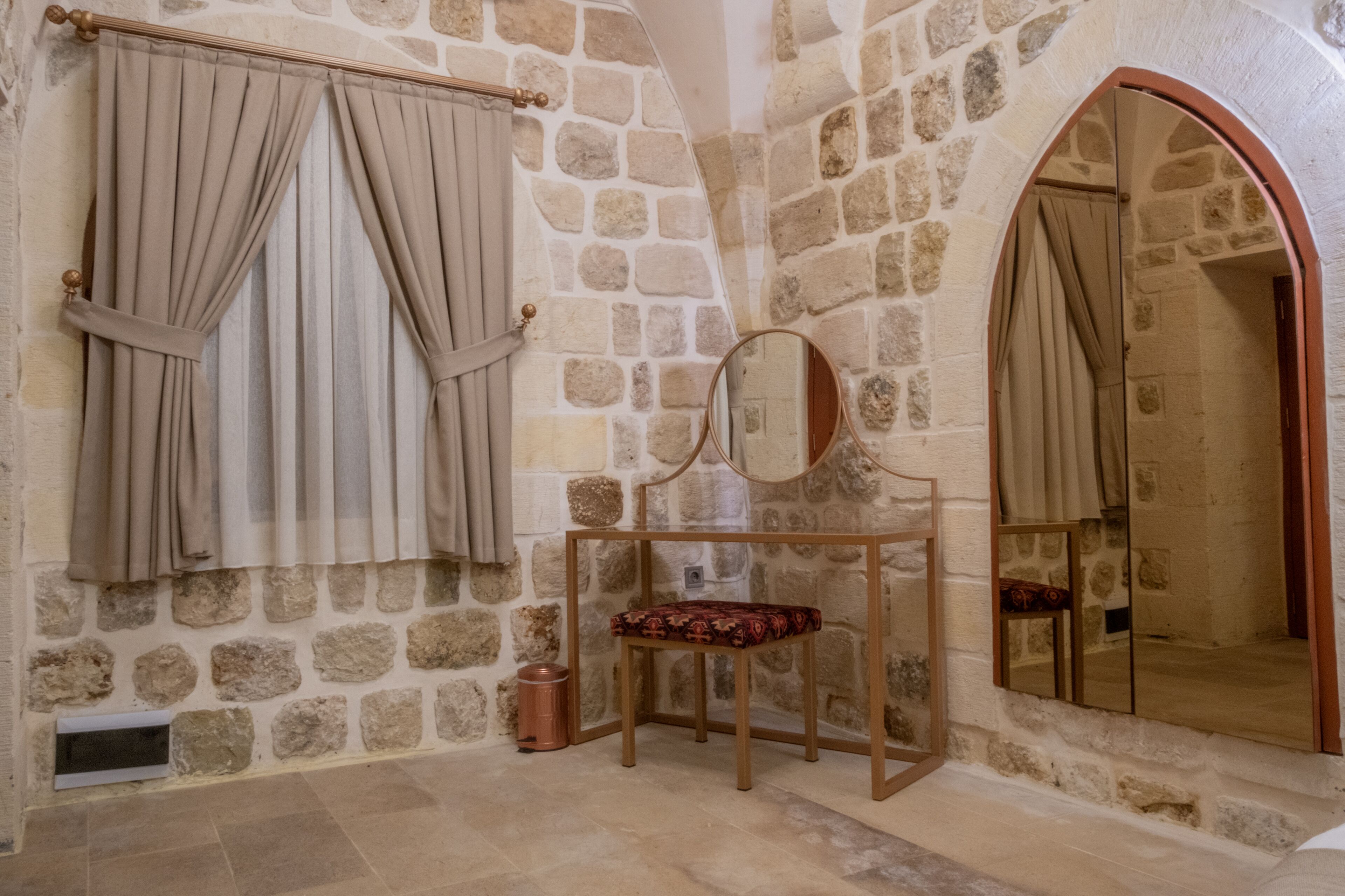 Une pièce aux murs de pierre, avec un banc, un miroir et une fenêtre ornée de rideaux.