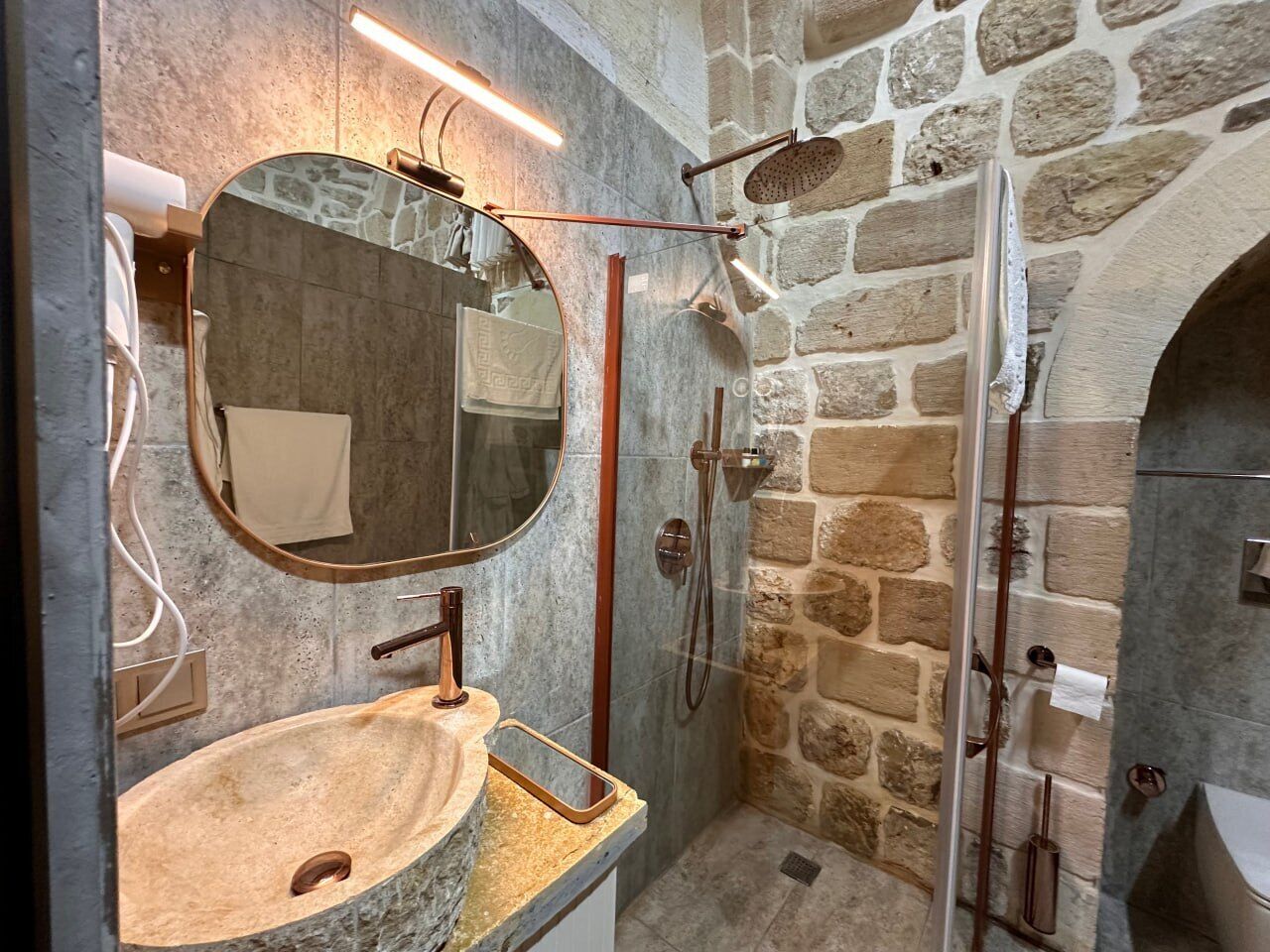 Un baño con pared de piedra, espejo redondo, lavabo y zona de ducha.