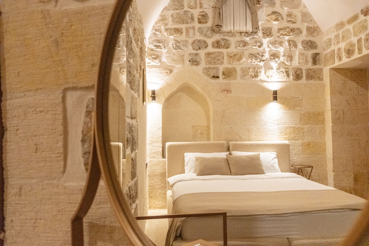 A stone-walled bedroom with a bed centered in the room, a small table, and a mirror reflecting part of the room.