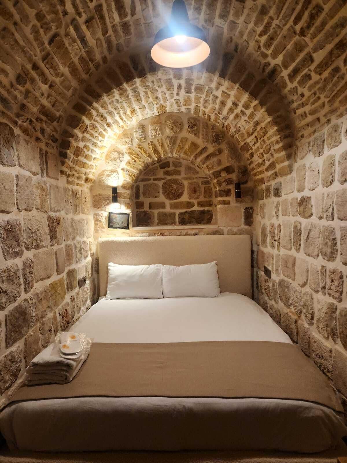 Una habitación con paredes de piedra, una cama, una mesita de noche y una lámpara de pared.