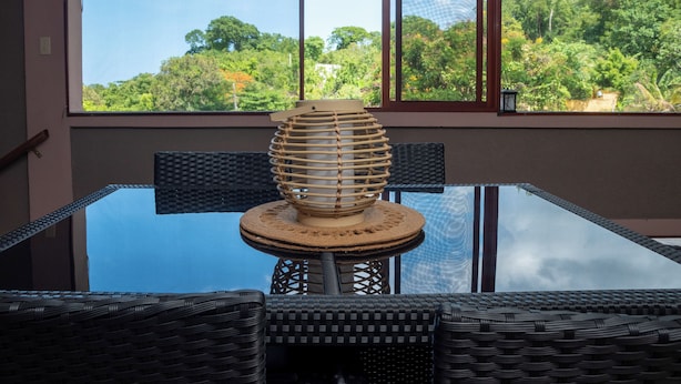 Een glazen tafel met een geweven stoel en een decoratief houten voorwerp, voor een raam met uitzicht op bomen en gebouwen.