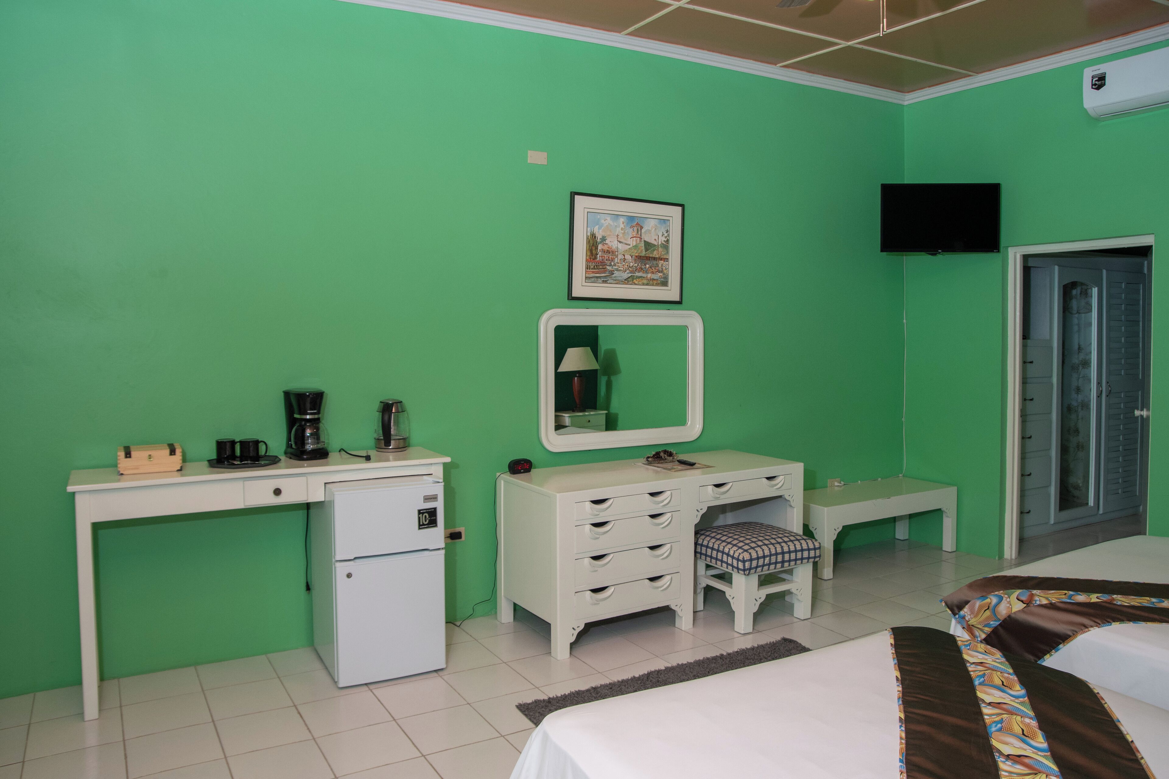 A room with a green wall, a white desk with a computer, a small refrigerator, a bed with a patterned bedspread, a mirror, and a television on the wall.
