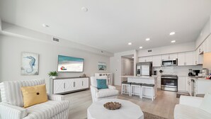 A modern living room with a sofa, coffee table, and a TV. Adjacent is a kitchen with white cabinets, a dining area, and a kitchen island. - BEACHFRONT PROPERTY, OUTDOOR POOL, GREAT LOCATION! (Gulf Shores)
