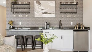 A modern kitchen with two black metal shelves, a stainless steel refrigerator, and a white countertop. There are two bar stools and a vase with green plants on the counter.