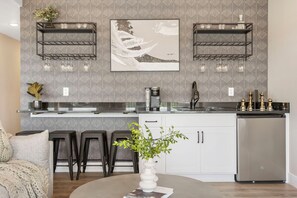 A modern kitchen with two black metal shelves, a stainless steel refrigerator, and a white countertop. There are two bar stools and a vase with green plants on the counter.