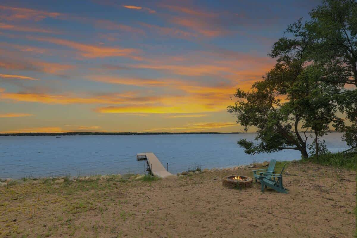 Un cadre idyllique au bord du lac, avec un foyer et des chaises, un chemin en béton menant à l’eau et un arbre offrant de l’ombre.