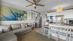 A modern kitchen with white cabinets, a dining area with white chairs, a ceiling fan, and a large abstract painting on the wall. - 3rd Floor Condo w/Amazing Ocean Views! Pool & Pier Access! In Heart of IOP! *Unit 321* (Isle Of Palms)