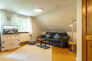 Ein gemütliches Wohnzimmer mit Fernseher, Sofa, Hocker und einer Stehlampe.