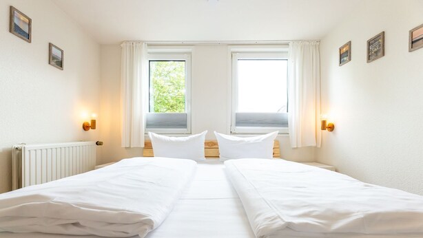 Habitación de hotel con dos camas individuales, un radiador, dos ventanas con cortinas e iluminación en la pared.