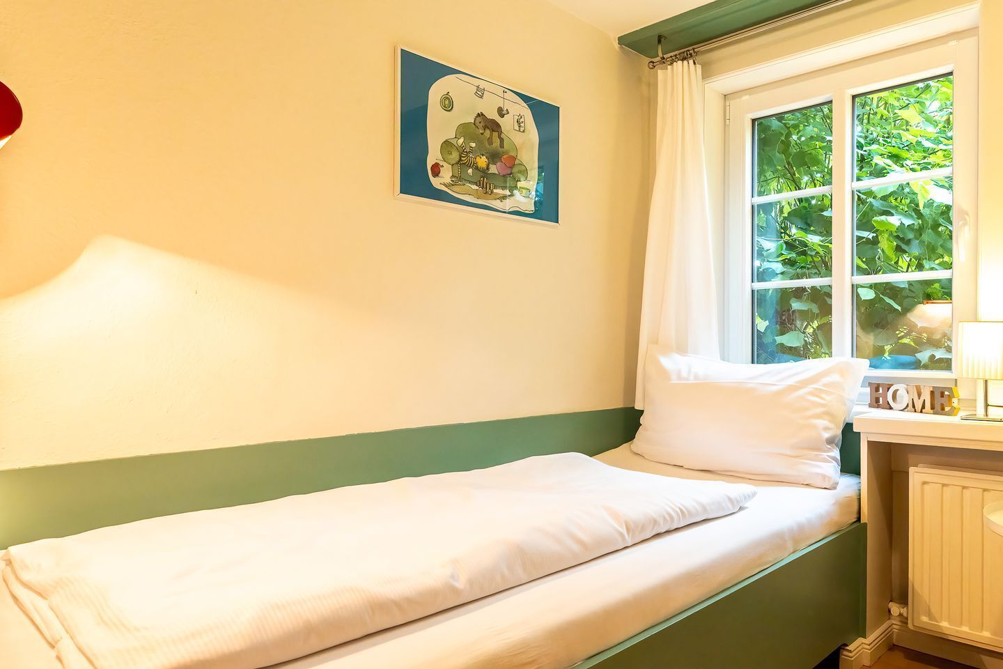 A single bed with white bedding, a green-painted wall, a window with a view of greenery, and a framed picture above the bed.