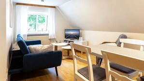 Ein gemütliches Wohnzimmer mit einer Couch, einem Fernseher, einem Esstisch mit Stühlen und einem Fenster mit Blick ins Grüne.