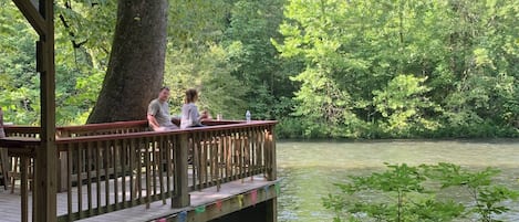 A wooden deck with a railing overlooking a body of water, surrounded by dense greenery.