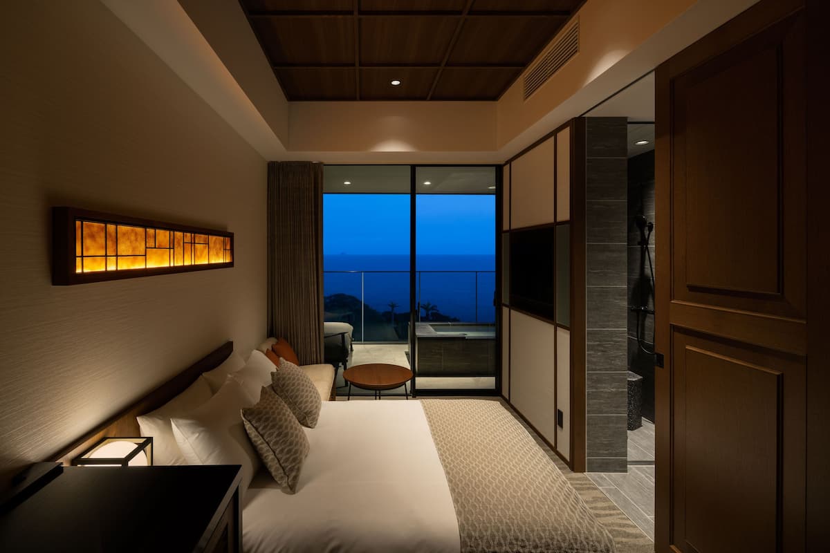A modern hotel room with a large bed, a small table, a TV, and a balcony with a view.