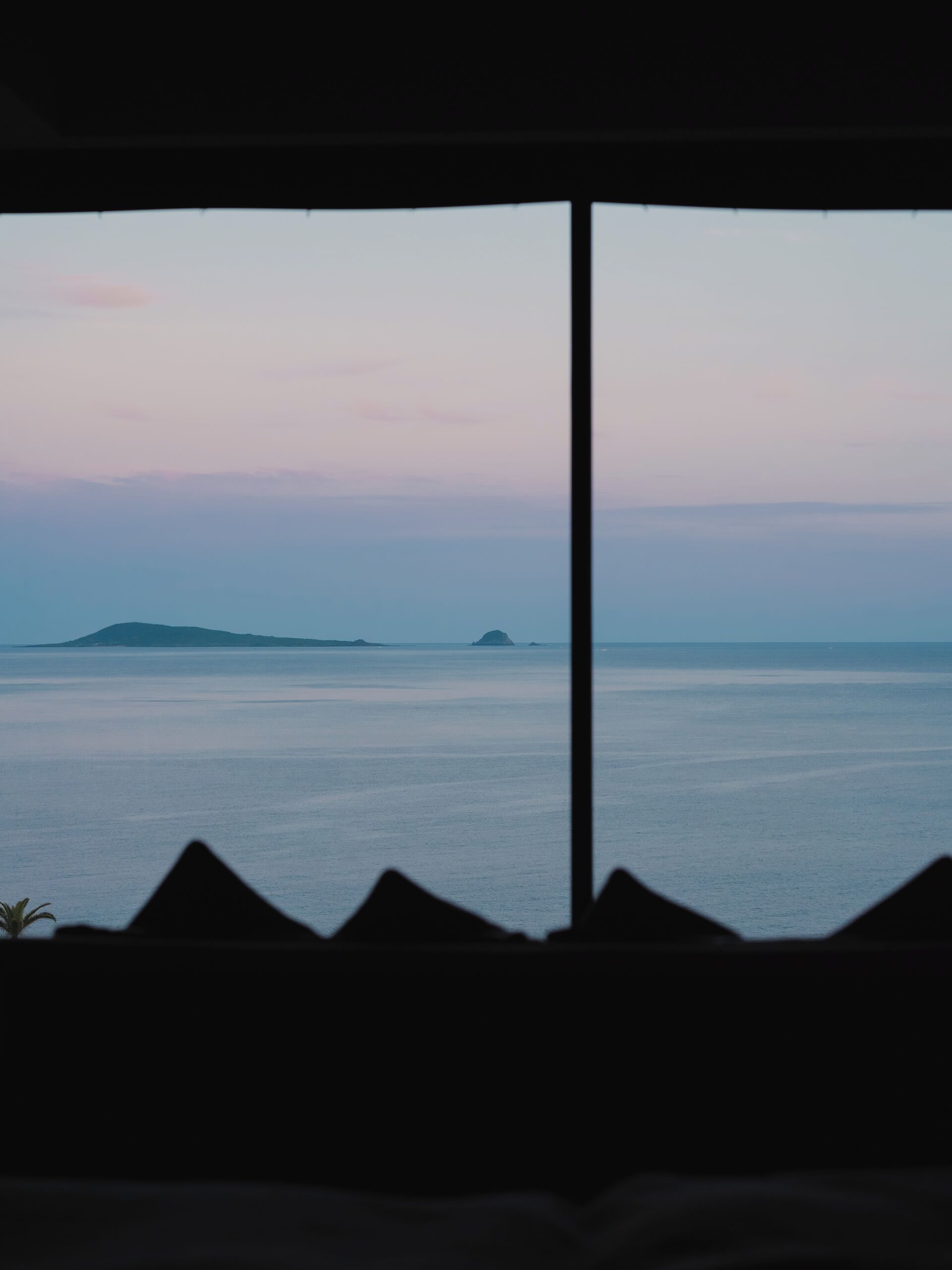 Vista a través de una ventana, con la silueta de una montaña al frente y un mar tranquilo con islas a lo lejos.