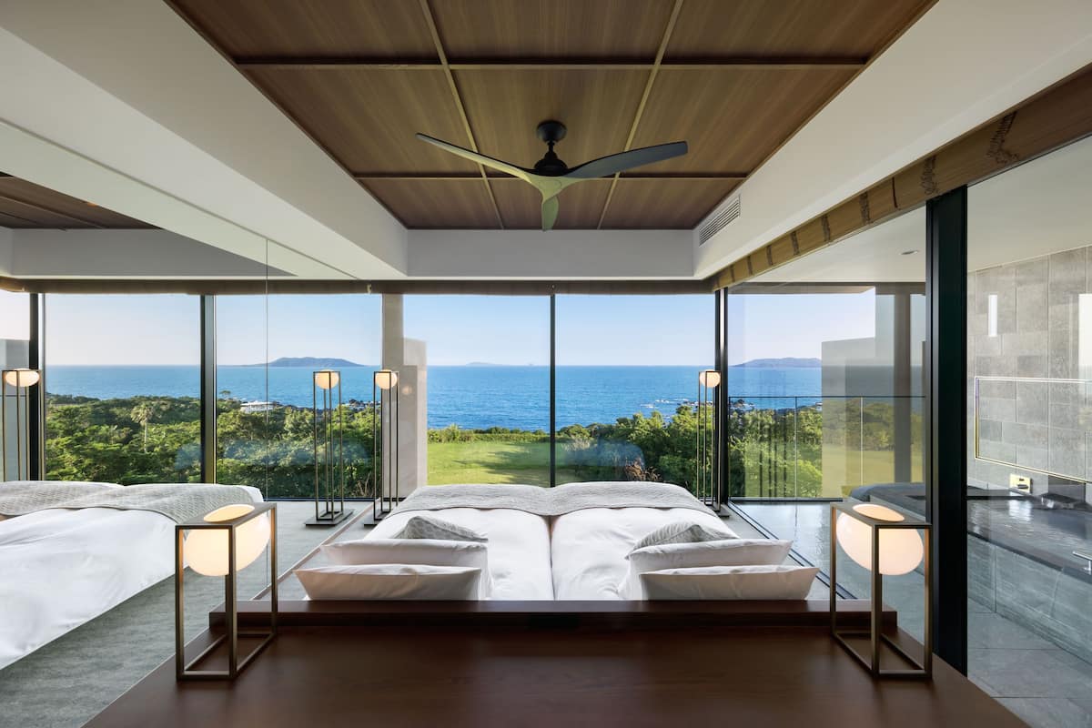 A modern bedroom with a large bed, two bedside tables, and a ceiling fan. It features large windows with a view of the sea and greenery.