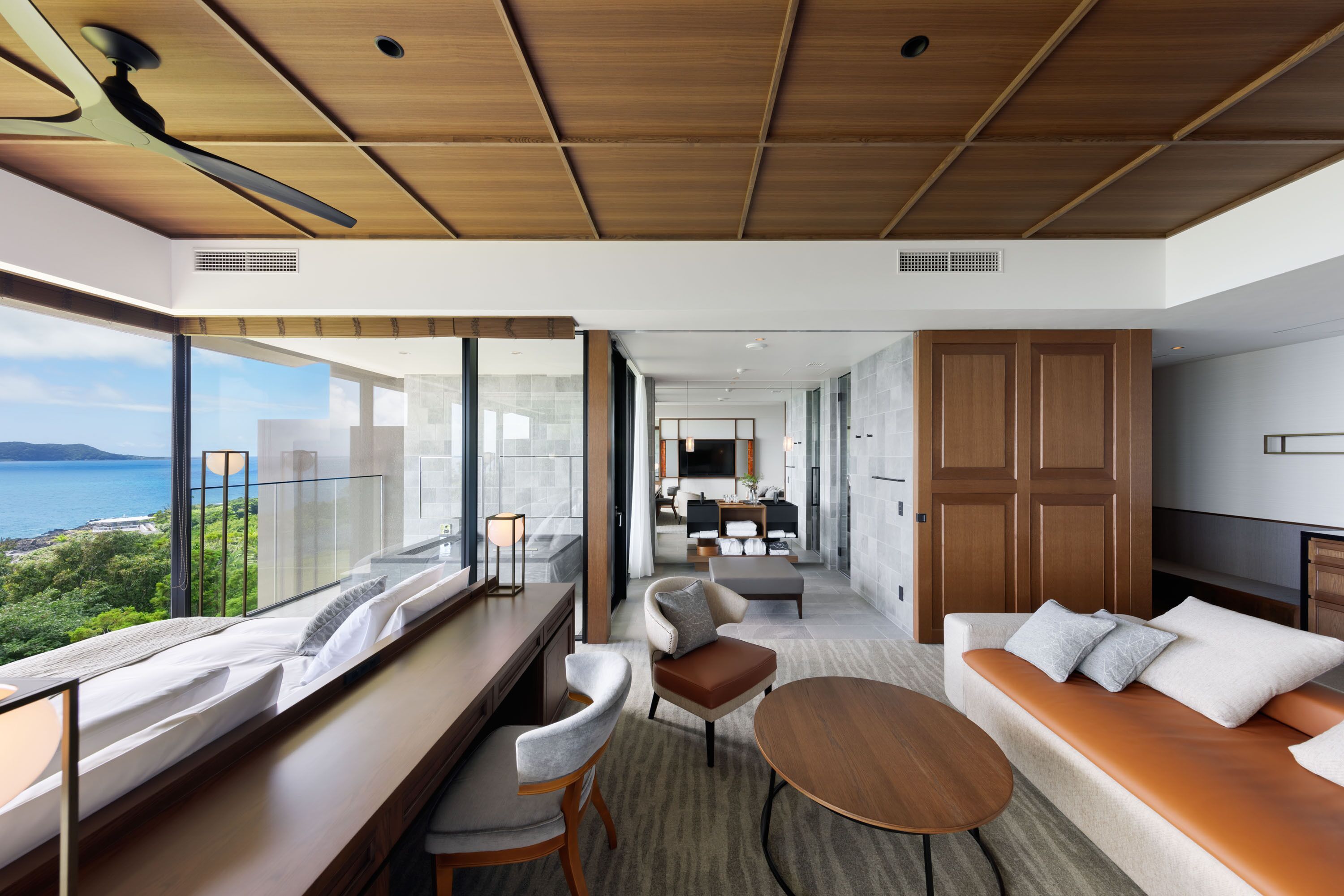 A modern hotel room with a large window, a sofa, a dining table, and a view of the sea.
