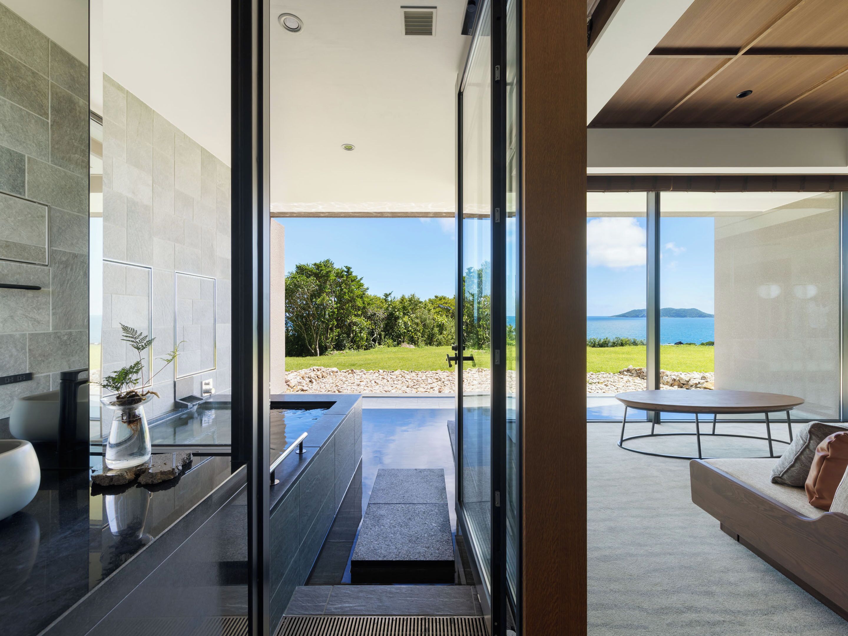 A modern interior with a glass wall leading to a balcony with a view of the sea and greenery.
