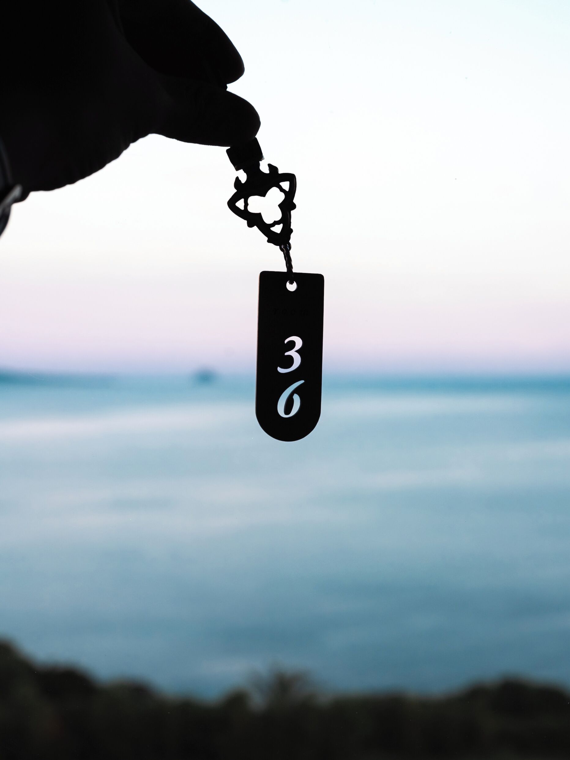 A silhouette of a person holding a keychain with the number 36 against a blurred background of a body of water and sky.