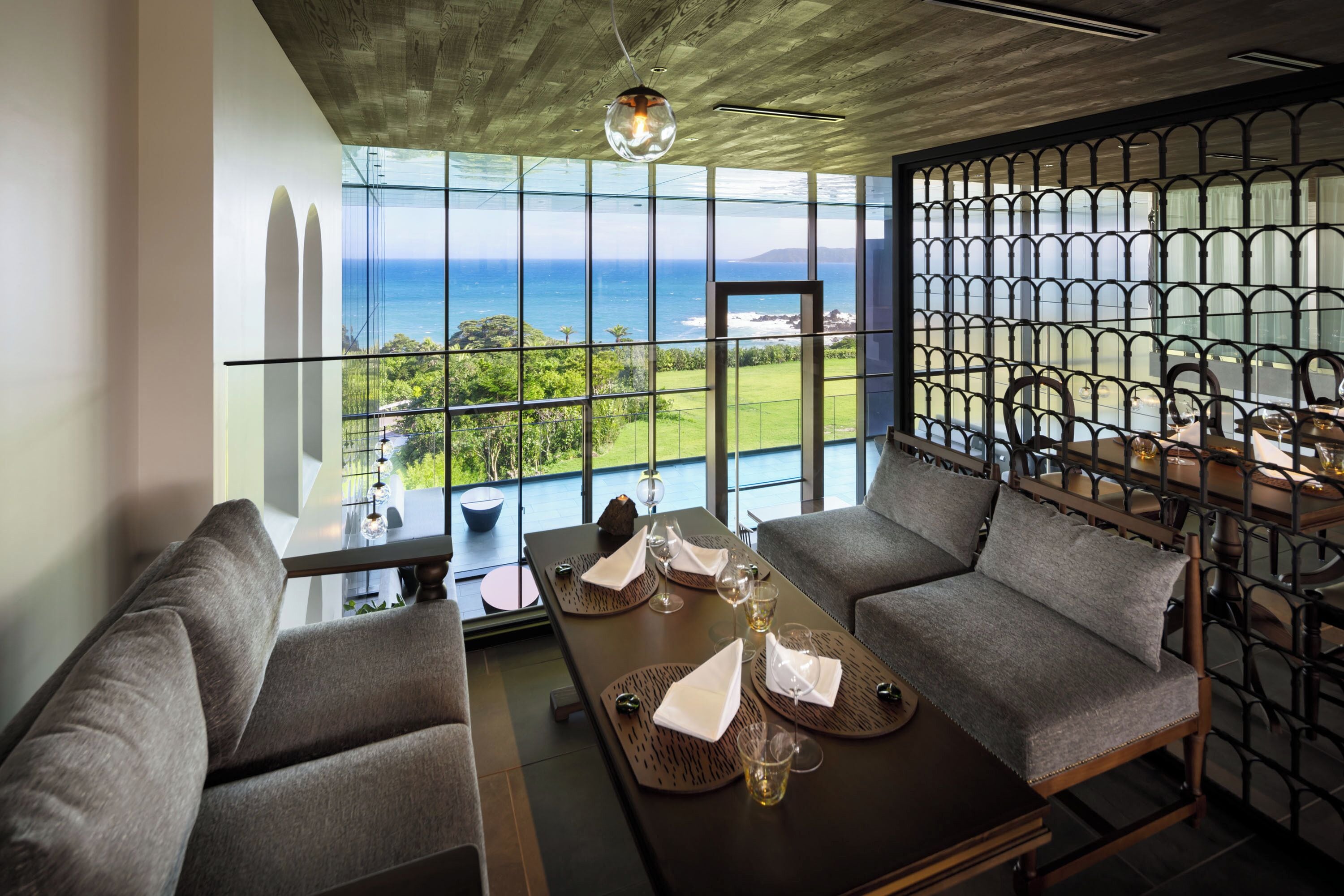 A modern dining area with a view of the ocean, featuring a long table with chairs, a sofa, and a decorative railing.