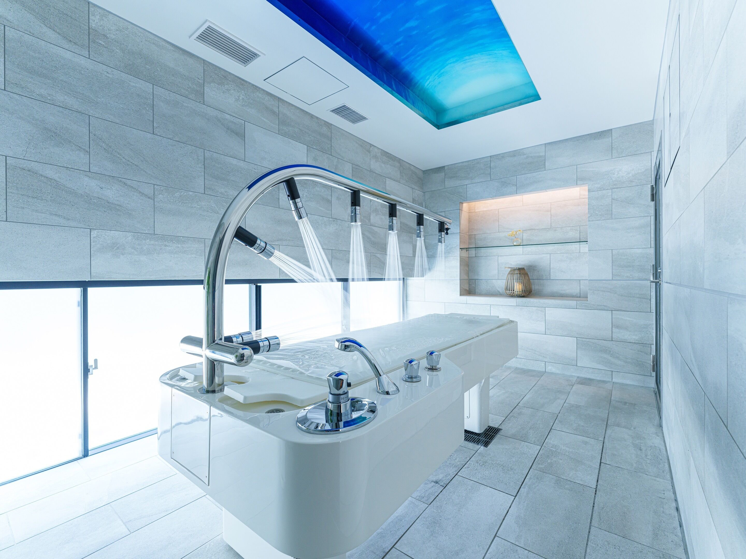 A modern bathroom with a large bathtub, multiple faucets, and a glass shelf with decorative items.