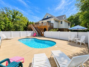 A backyard with a swimming pool, white deck chairs, a wooden deck, and a white fence.