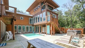 Een modern meerlaags huis met een groot buitenzwembad, een houten terras en een zitgedeelte buiten.