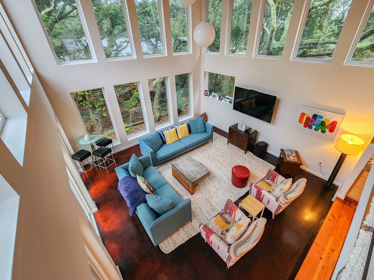 A spacious living room with a large blue sofa, a coffee table, and a TV mounted on the wall. There are multiple windows providing a view of the outdoors.