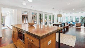 Een ruime keuken met een groot eiland, houten kastjes en een ingebouwde oven. Er is een zithoek met een tafel en stoelen, en een uitzicht op de omgeving door grote ramen.