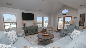A spacious living room with a large sofa, a coffee table, and a television. There is a balcony with a railing and a view of the outside. - The Penny Swan - Wake up to beautiful ocean views from this oceanside 7BR home w/Private Pool! (Southern Shores)