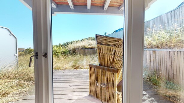 Een houten terras leidt naar een overdekt buitenverblijf met een rieten stoel.