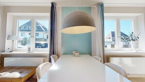 Een eetgedeelte met een witte tafel, een grijze hanglamp en een uitzicht op gebouwen door grote ramen.