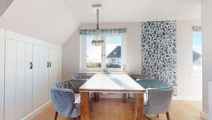 Un espace repas comprenant une longue table, six chaises, une fenêtre donnant sur un paysage agréable et un mur décoré de motifs.