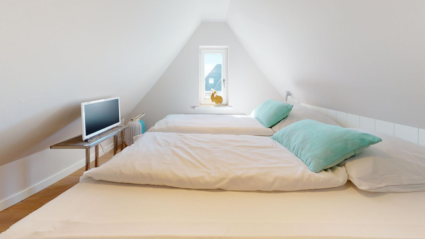 Une petite chambre mansardée de forme triangulaire, avec deux lits, une télévision et une fenêtre donnant sur une maison.