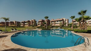 Ein Swimmingpool mit glasklarer, blauer Wasseroberfläche, umgeben von Liegestühlen und Palmen, mit modernen Gebäuden im Hintergrund.