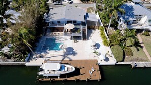 Vista aérea de una casa con una amplia piscina, un barco atracado en un muelle de madera y un jardín cuidado con palmeras.