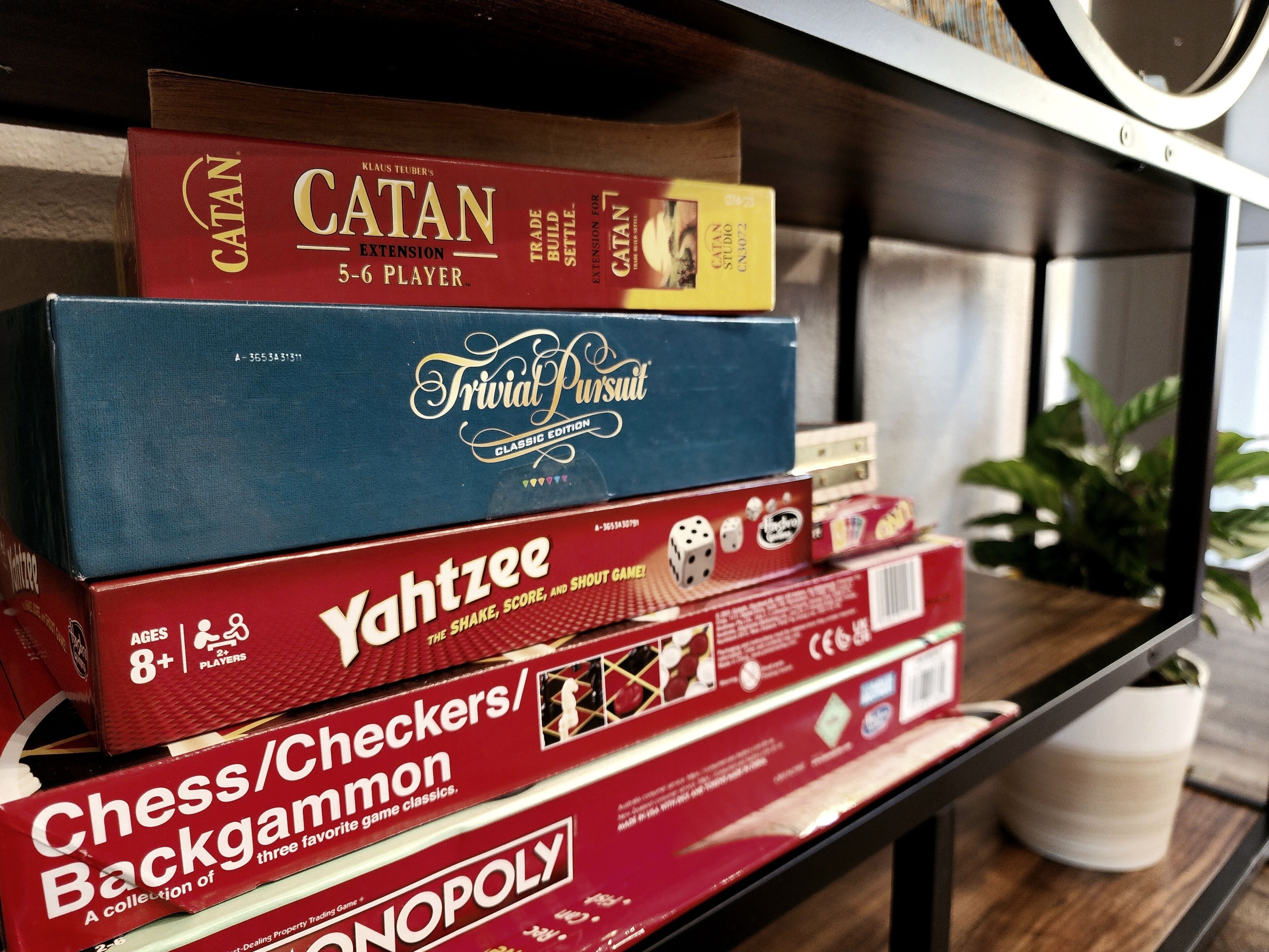 A shelf with various board games including Catan, Trivial Pursuit, Yahtzee, and Chess/Checkers/Backgammon.