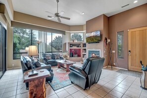 Un salón amplio con sofás de cuero, chimenea, televisor de pantalla plana y ventanas grandes con vistas a árboles.