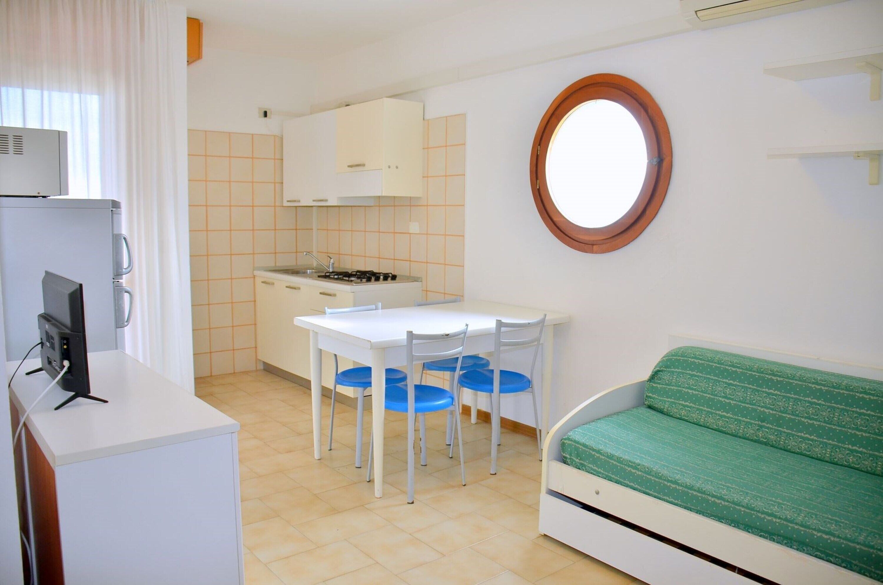 A compact kitchen with a dining table and chairs, a small refrigerator, and a round window.