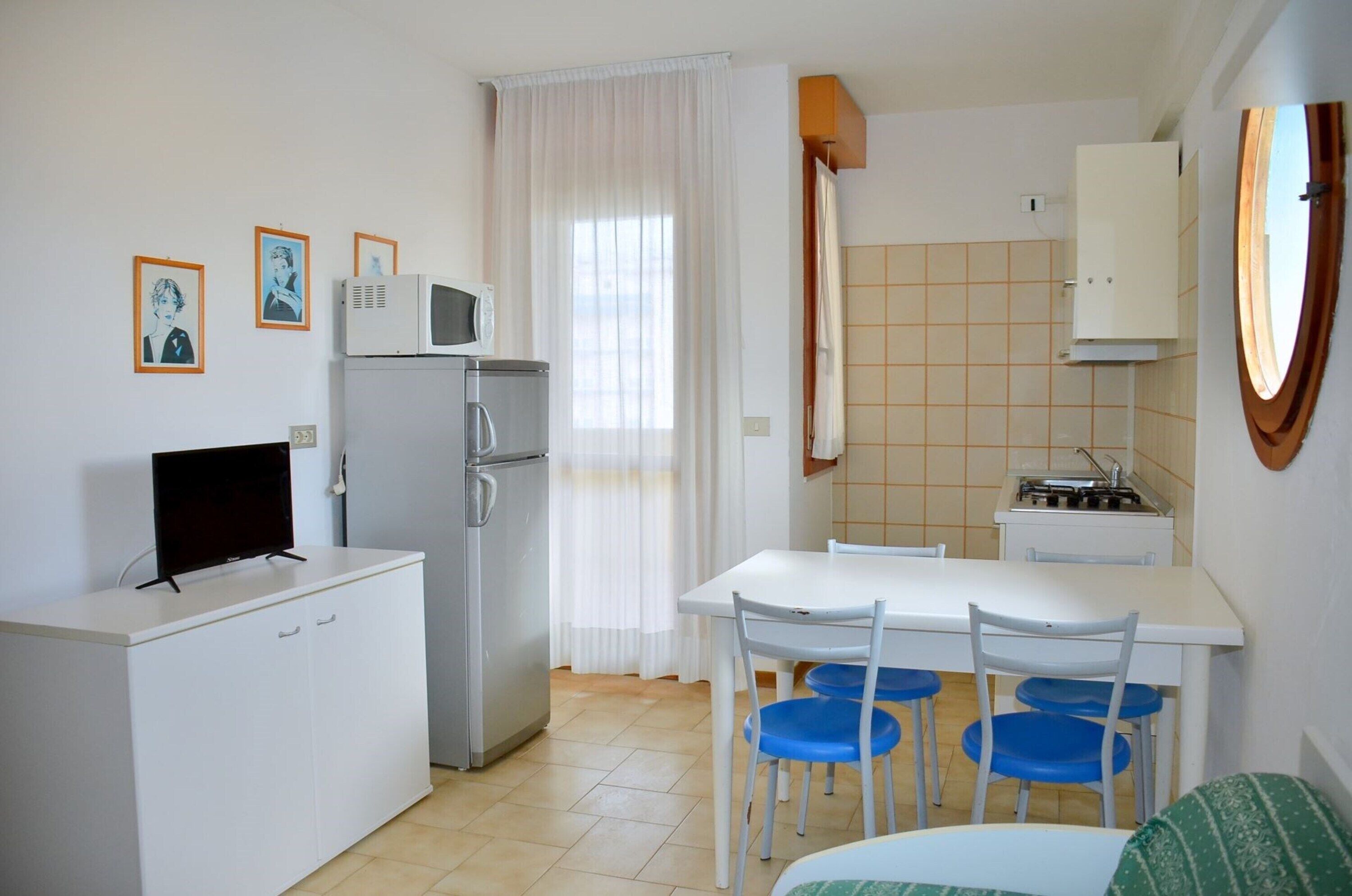 A compact kitchen with a refrigerator, microwave, and stove. There is a dining table with blue chairs and a white cabinet.