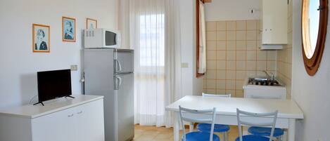 A compact kitchen with a refrigerator, microwave, and stove. There is a dining table with blue chairs and a white cabinet.