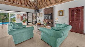 A spacious living room with a high wooden ceiling, a large TV, and a pool visible through the glass doors. - Forever Summer Gorgeous 4BDR Villa (Runaway Bay)