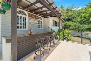 A bar area with a wooden counter and three stools, a sign reading "Gilmour's Bar," and a view of greenery and a fence. - Forever Summer Gorgeous 4BDR Villa (Runaway Bay)