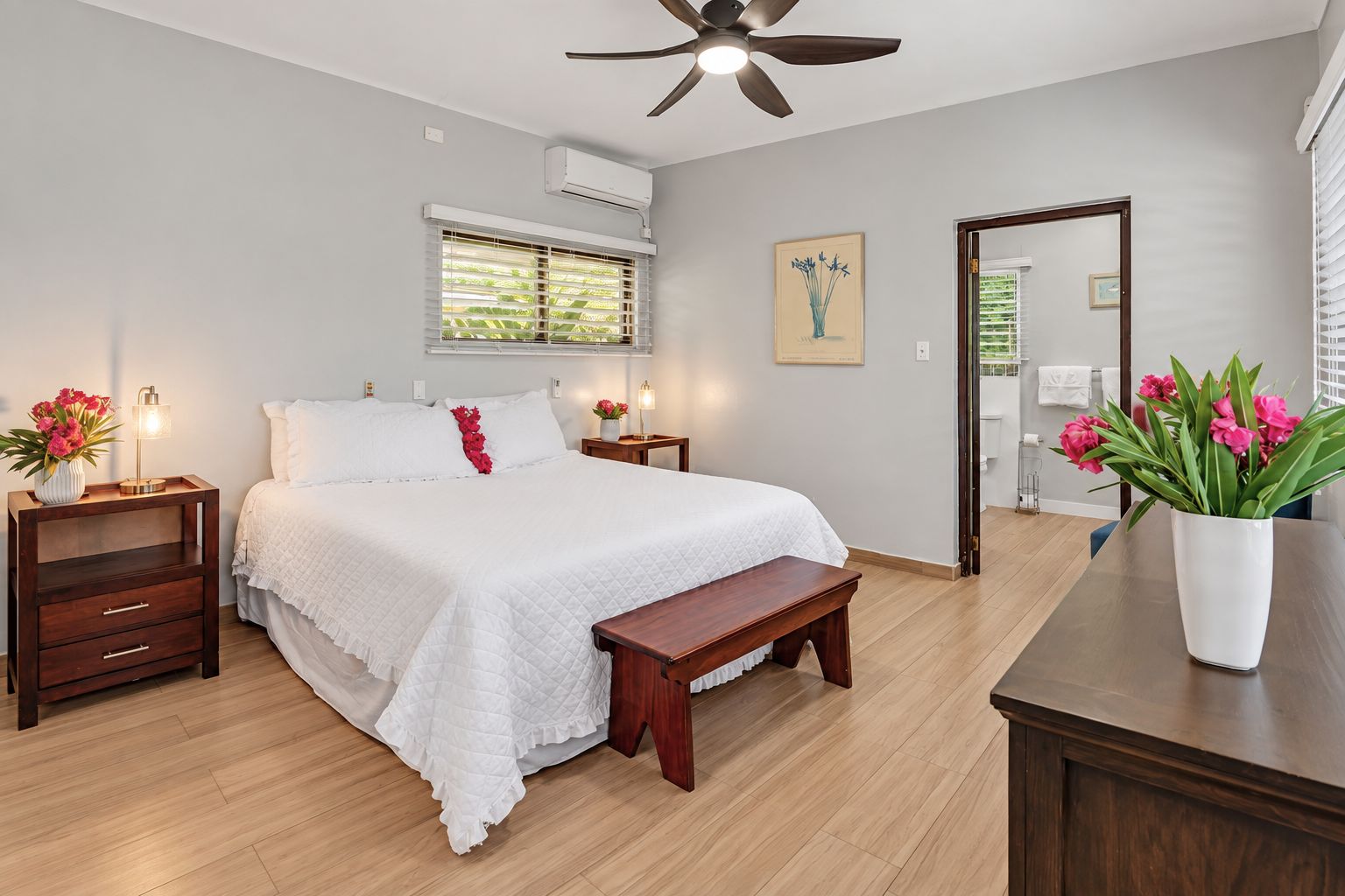 A bedroom with a bed, bedside tables, a bench, a ceiling fan, an air conditioning unit, and a window with blinds.