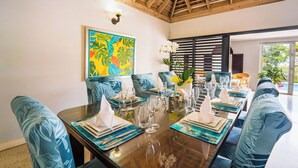 A dining area with a wooden table set for a meal, blue upholstered chairs, and a colorful tropical-themed painting on the wall. - Forever Summer Gorgeous 4BDR Villa (Runaway Bay)