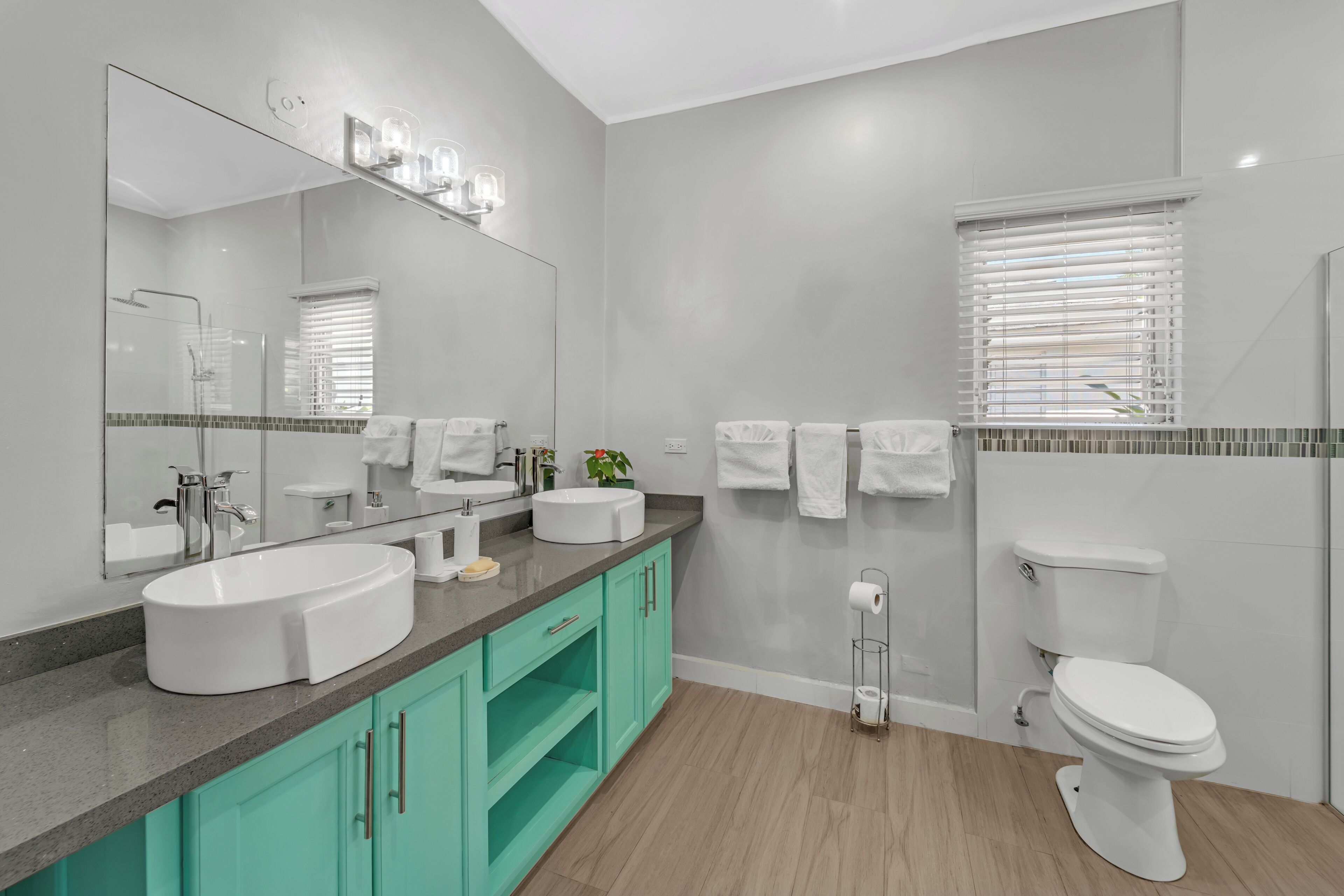 A modern bathroom with a large mirror, two sinks, a toilet, and a window with blinds.